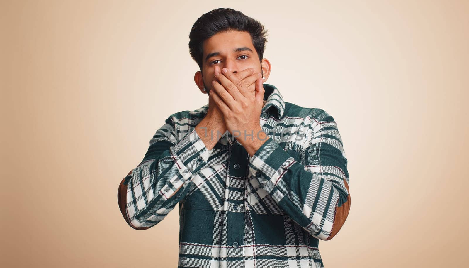 I will not say anyone. Frightened indian man closing mouth with hands, looking intimidated scared at camera, gestures no, refusing to tell terrible secret, unbelievable truth. Guy on beige background
