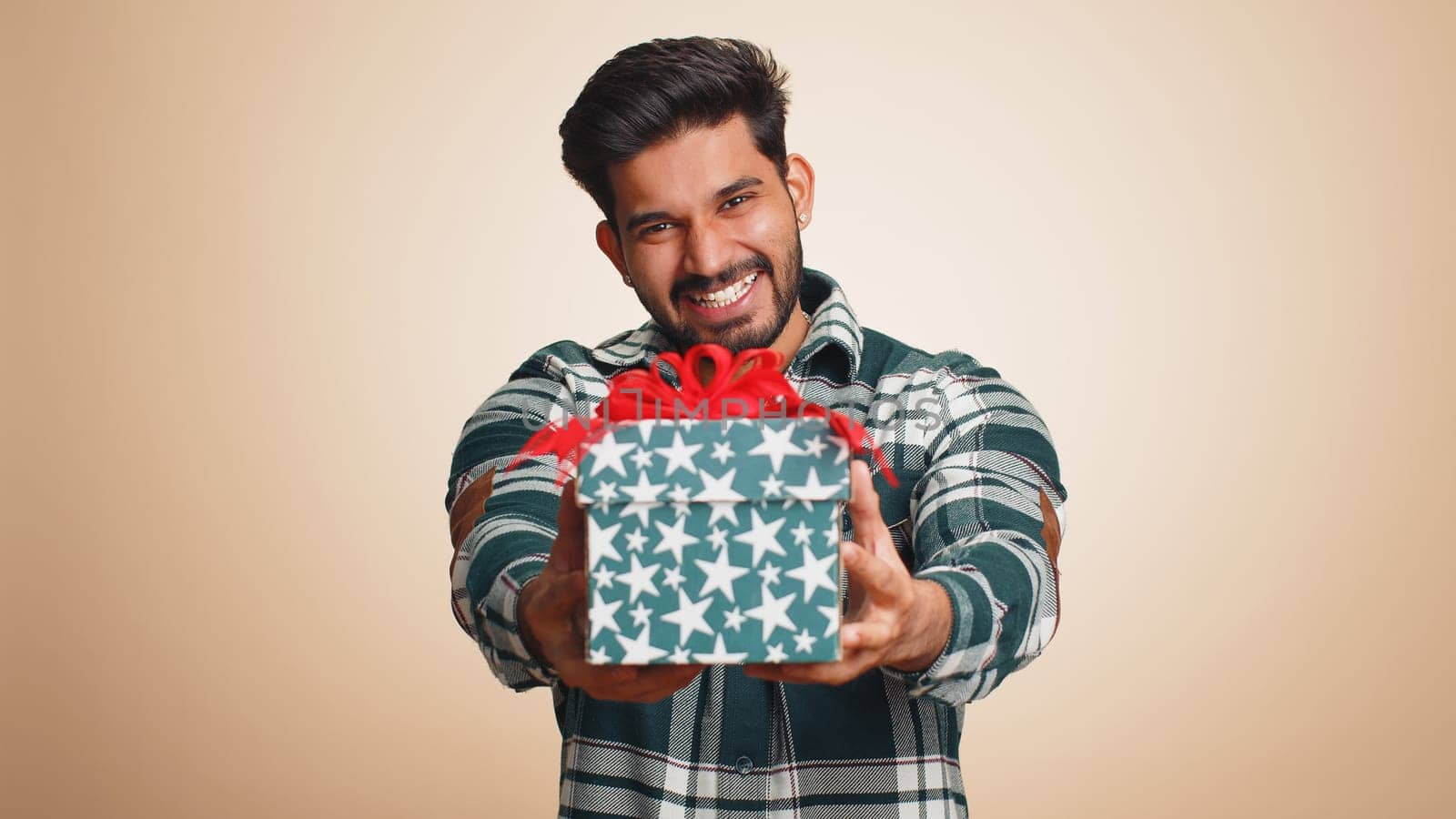 Positive smiling young indian man presenting birthday gift box stretches out hands, offer wrapped present career bonus, celebrating party, sale. Bearded hindu guy isolated on beige studio background