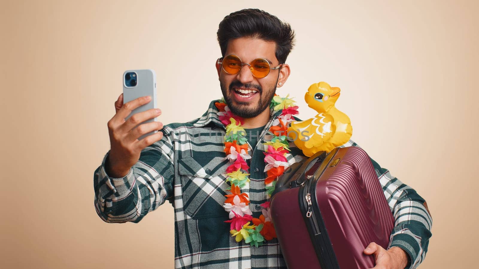 Young indian man traveler blogger in modern sunglasses with swimming ring, luggage taking selfie on smartphone with subscribers record make video call online. Hindu tourist guy on beige background