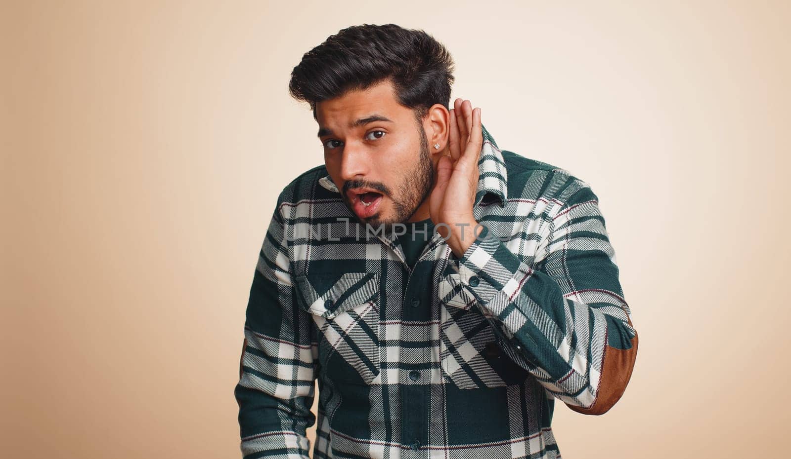 I cant hear you. What. Young indian man trying hear, looking confused, frowning, keeping arm near ear for louder voice, asking to repeat, to hear information, deafness. Hindu guy on beige background