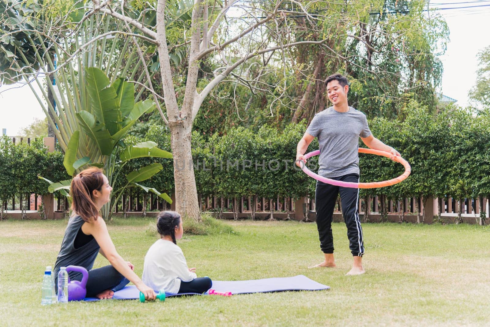 Asian young mother and child daughter practicing doing yoga exercises outdoors cheer father playing with hula hoops in nature a field garden park, family kid sport and exercises for healthy lifestyle