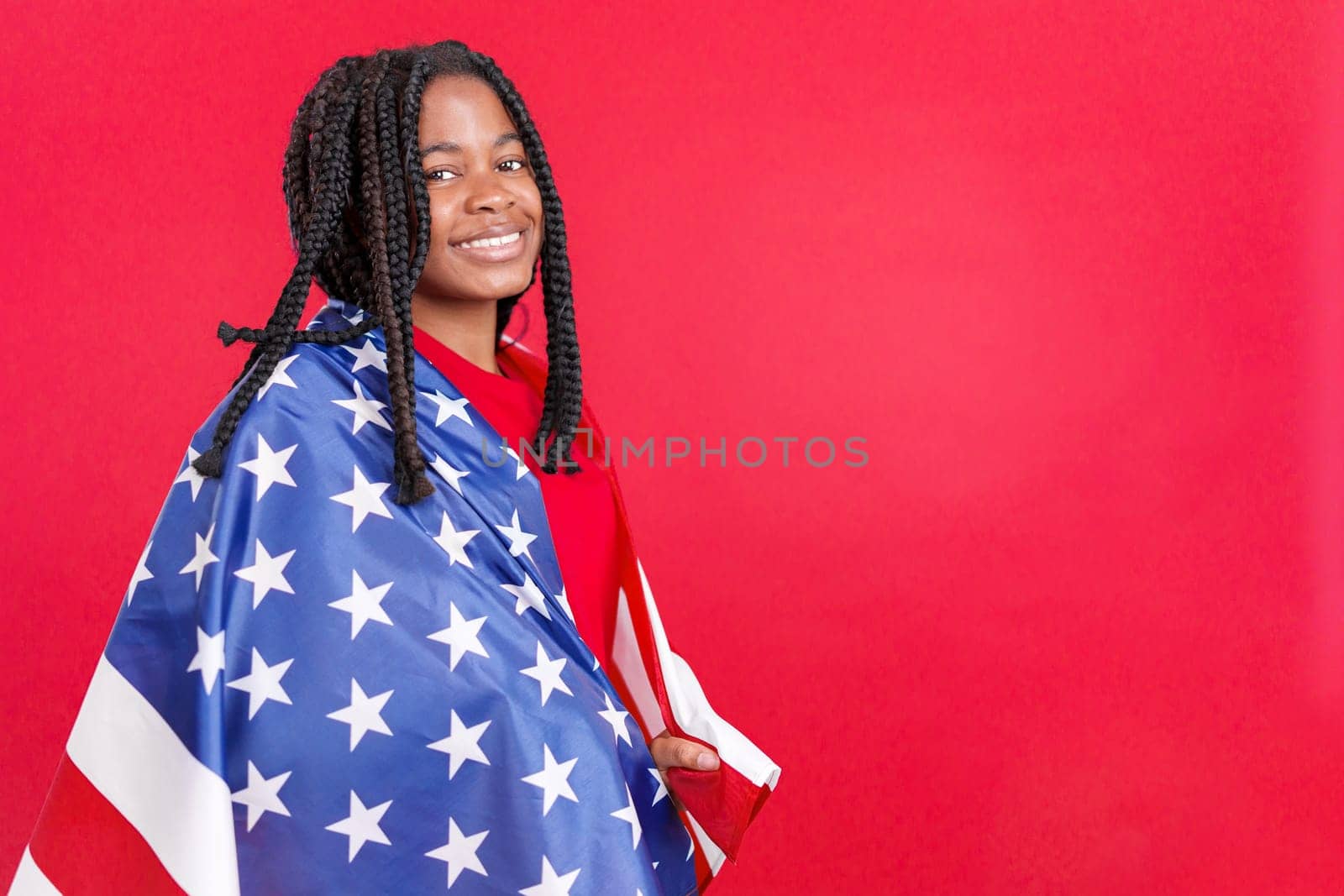 Happy african woman wrapped with a USA national flag in studio with red background