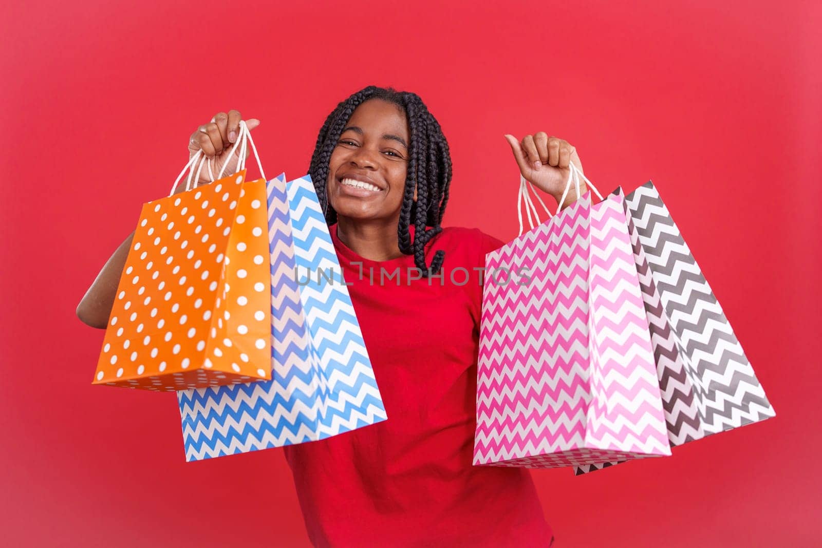 African woman raising many shopping bags while smiling at camera in studio with red background