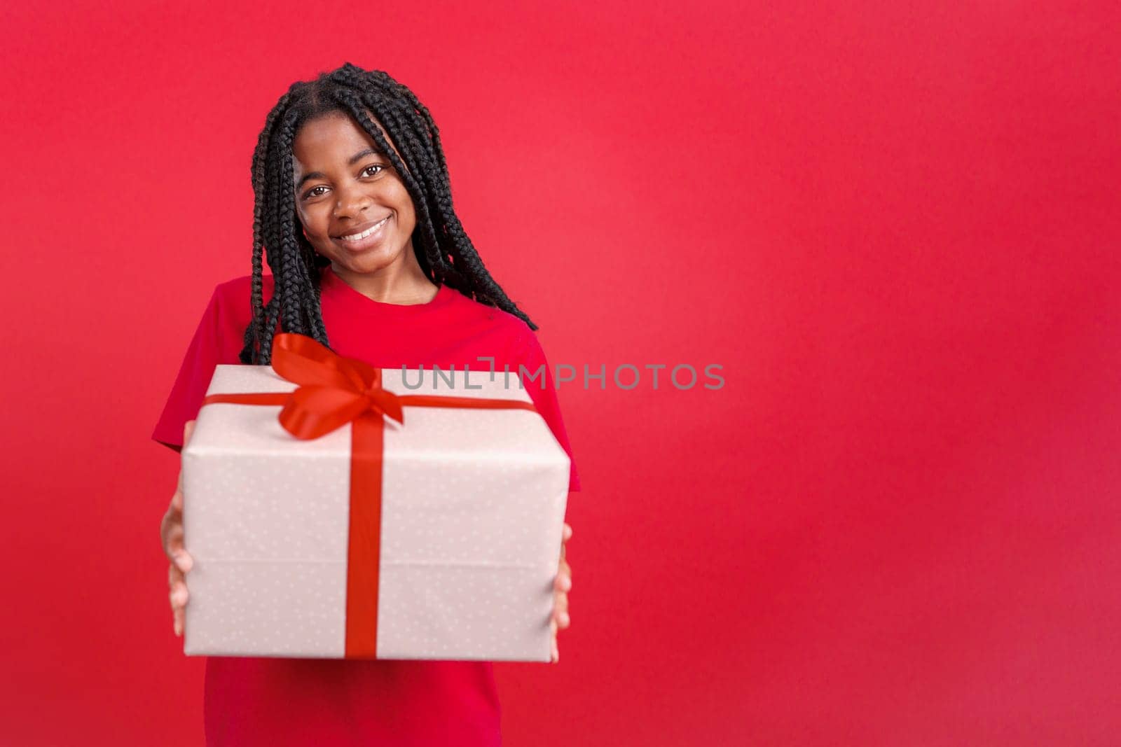 Happy african woman looking at the camera giving a present in studio with red background