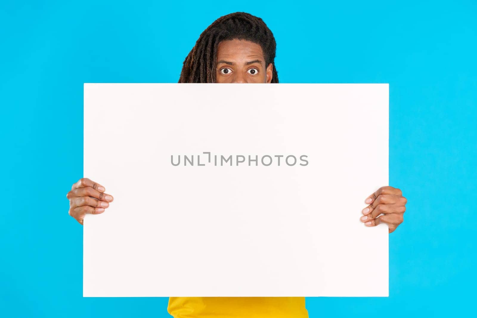 Surprised latin man with dreadlocks hiding behind a blank board in studio with blue background