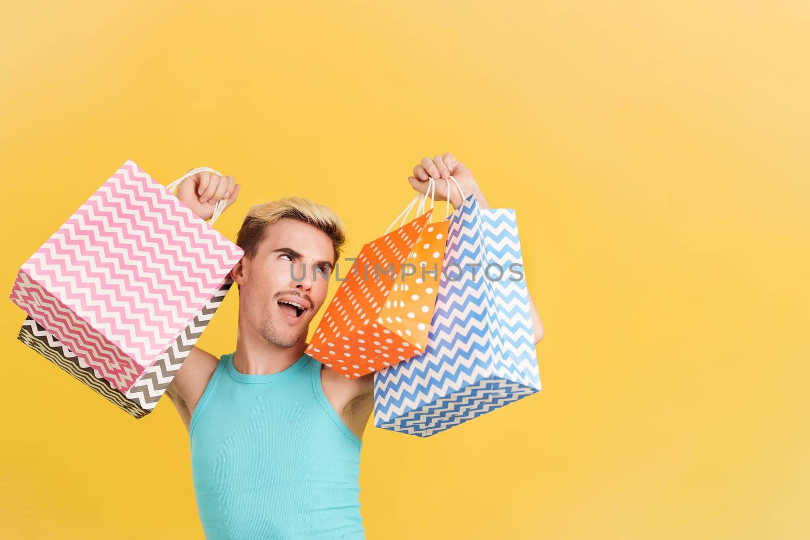 Cute gay man raising many shopping bags while smiling in studio with yellow background