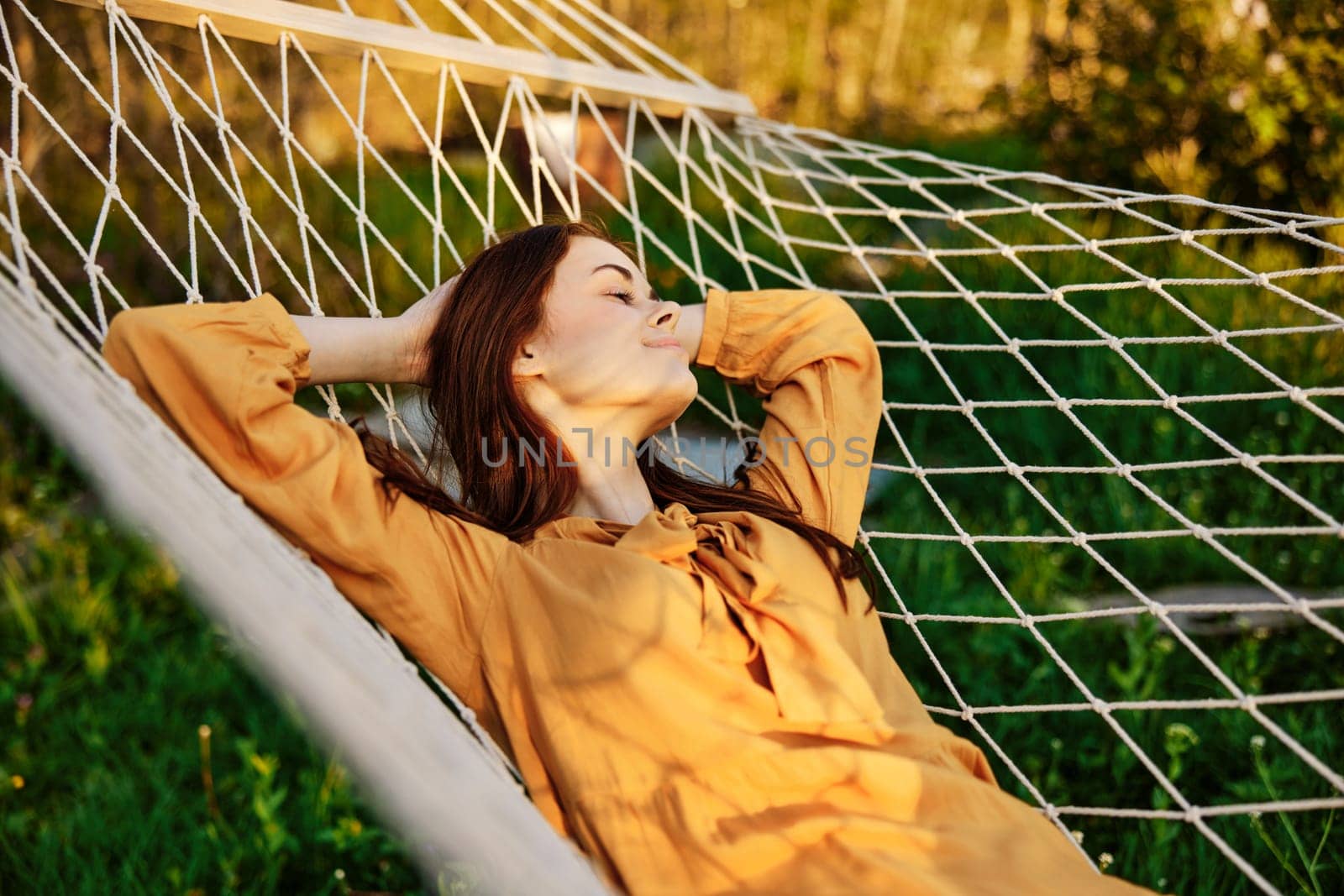 a happy woman is resting in a hammock with her eyes closed and her hands behind her head smiling happily enjoying the day by Vichizh
