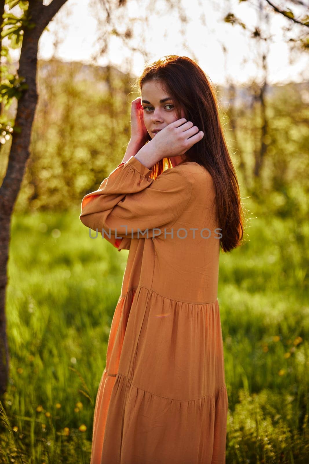 a sweet, thoughtful woman stands in nature near a tree in a long orange dress, illuminated from the back by the sunset rays of the sun and holding her hand near her face looks towards the camera. High quality photo