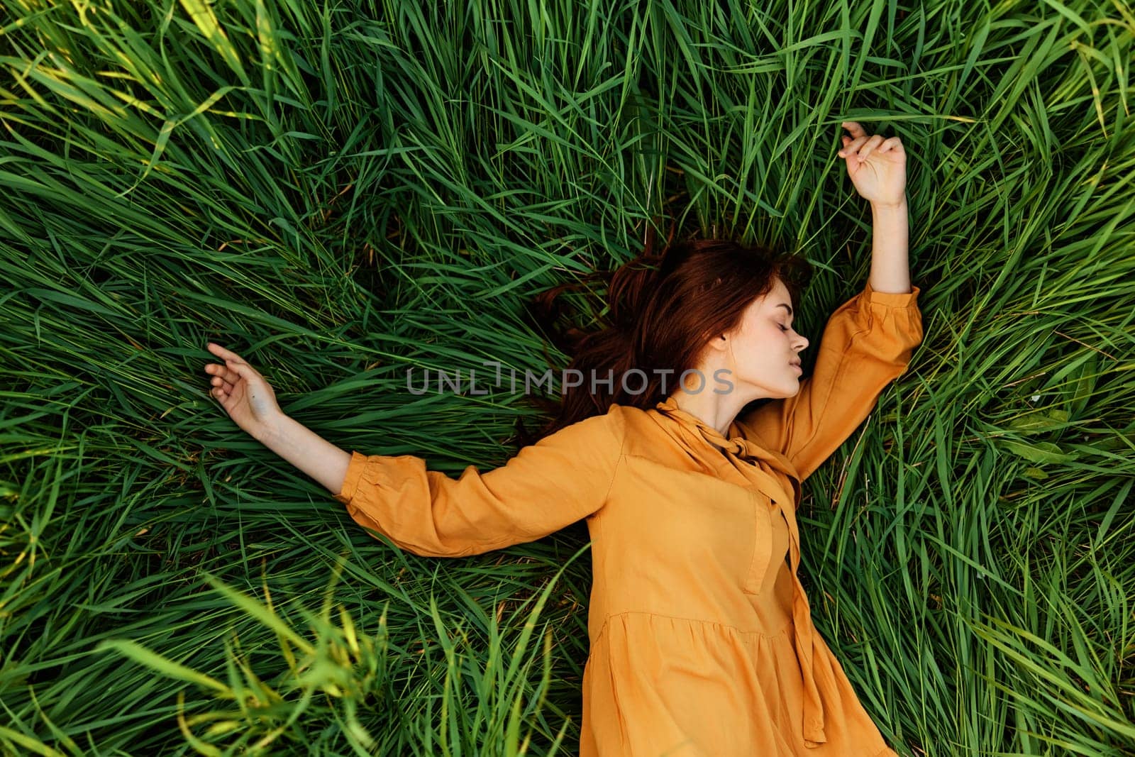 an elegant woman lies in the tall green grass in a long summer orange dress and with her arms outstretched enjoys nature and sunny weather with her eyes closed by Vichizh