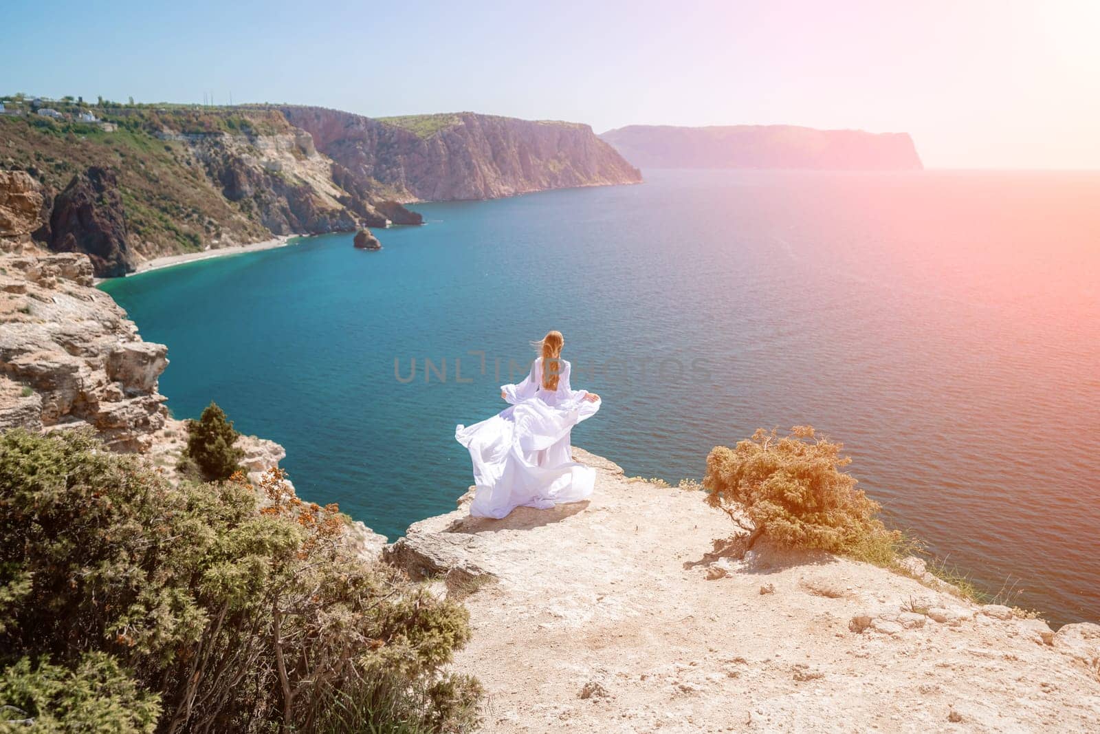 woman sea white dress. Blonde with long hair on a sunny seashore in a white flowing dress, rear view, silk fabric waving in the wind. Against the backdrop of the blue sky and mountains on the seashore. by Matiunina