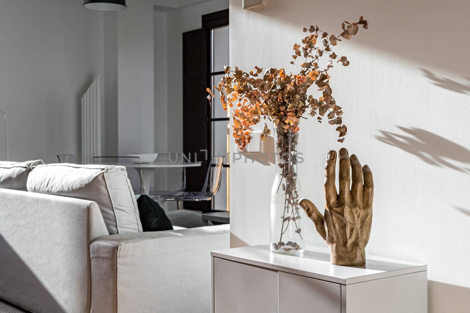Decorative hand sculpture and vase with flowers on small white cupboard in sunny interior of modern apartment.