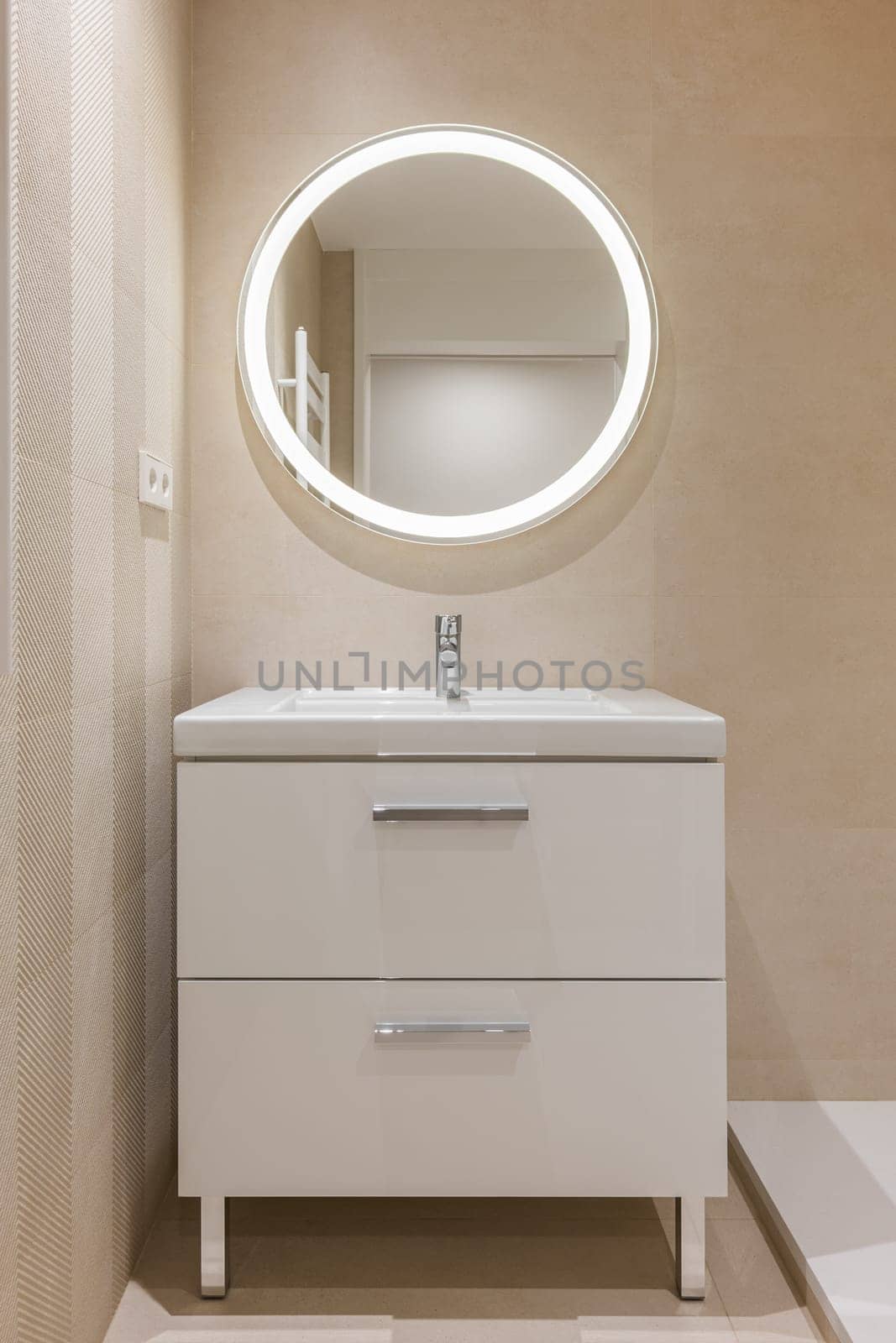 Modern bathroom with beige tiles, furniture and round large mirror with lighting.