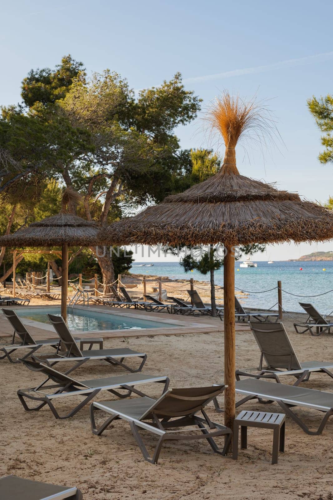 Sunbeds and thatched sunshades on the beach of a hotel with swimming pool and view to the sea on Ibiza island. Beautiful relax zone with the evening light. by apavlin