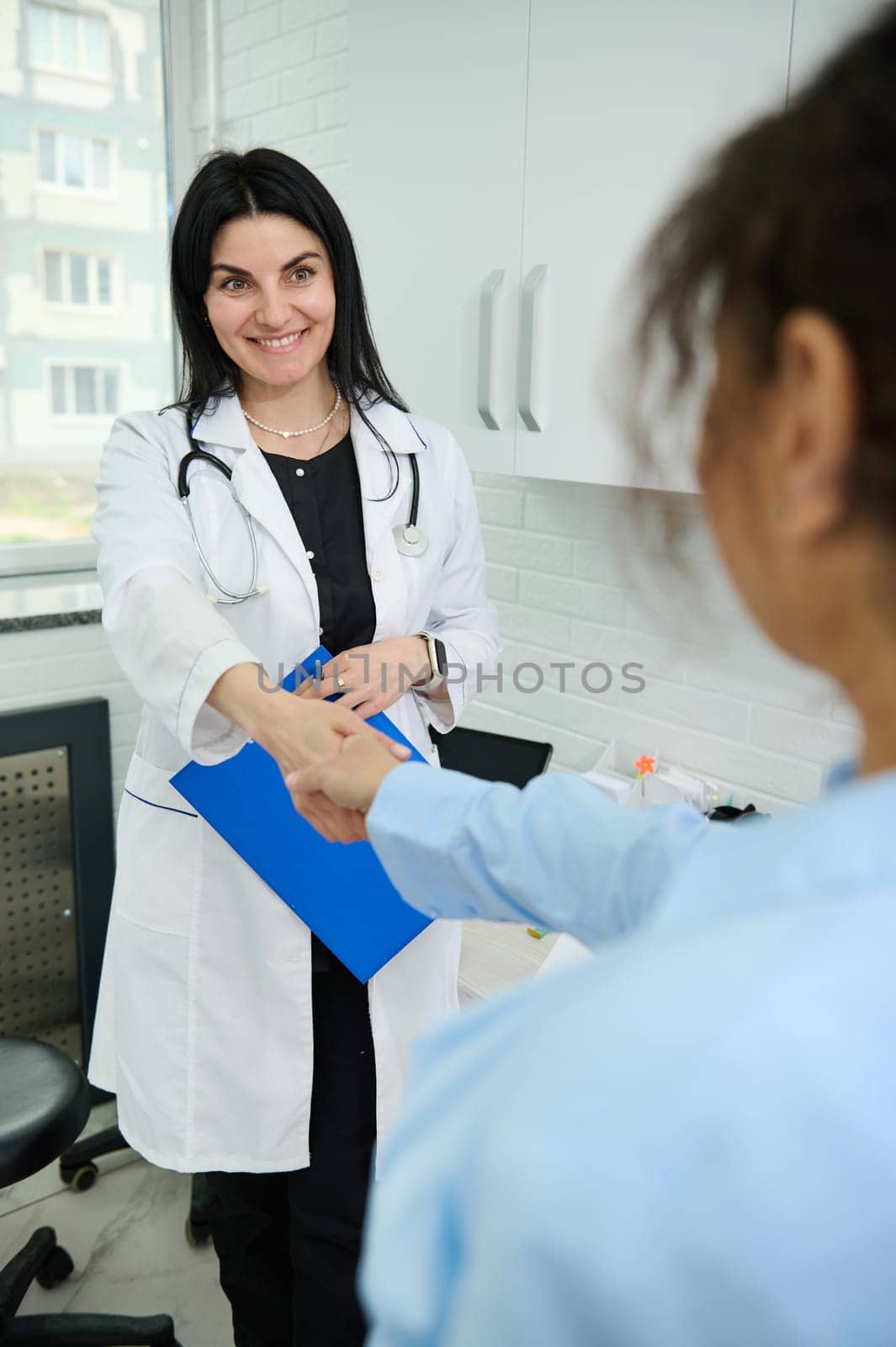Charming pleasant female doctor holding clipboard, smiling, shaking hand and welcoming a pregnant patient in her office by artgf