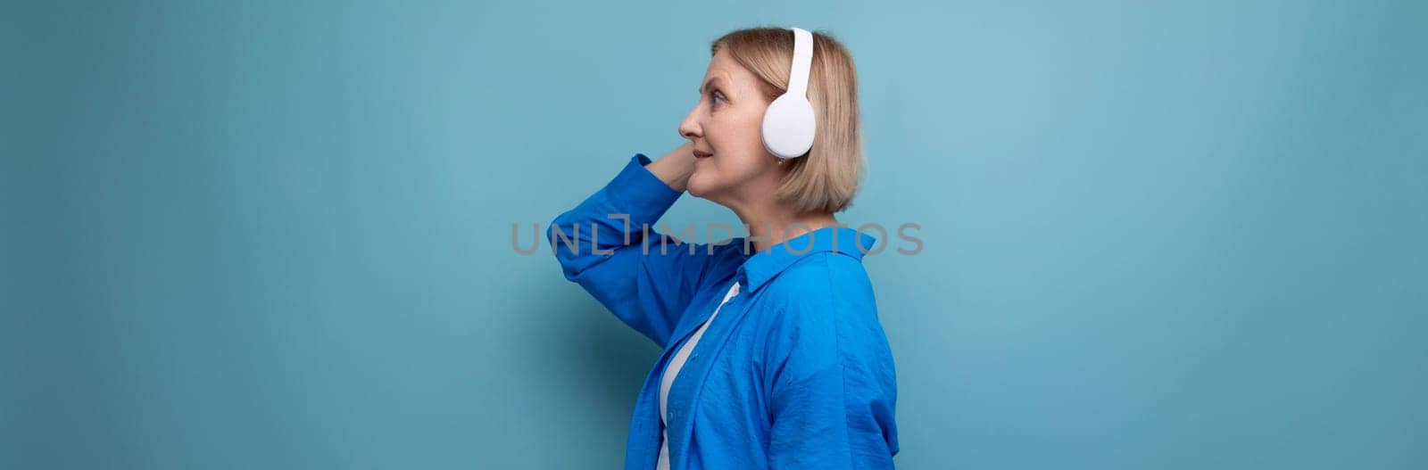 middle age business concept. mature woman listening to audiobook with headphones on blue background copy space.