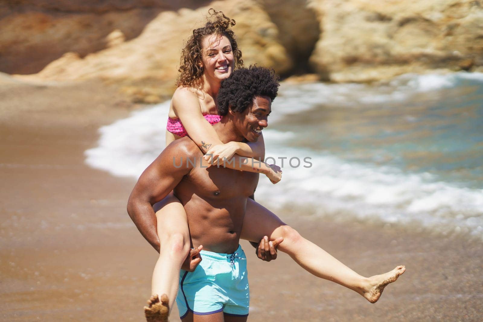 Happy young multiracial couple fooling around on sandy beach near waving ocean while enjoying summer vacation together during sunny weekend