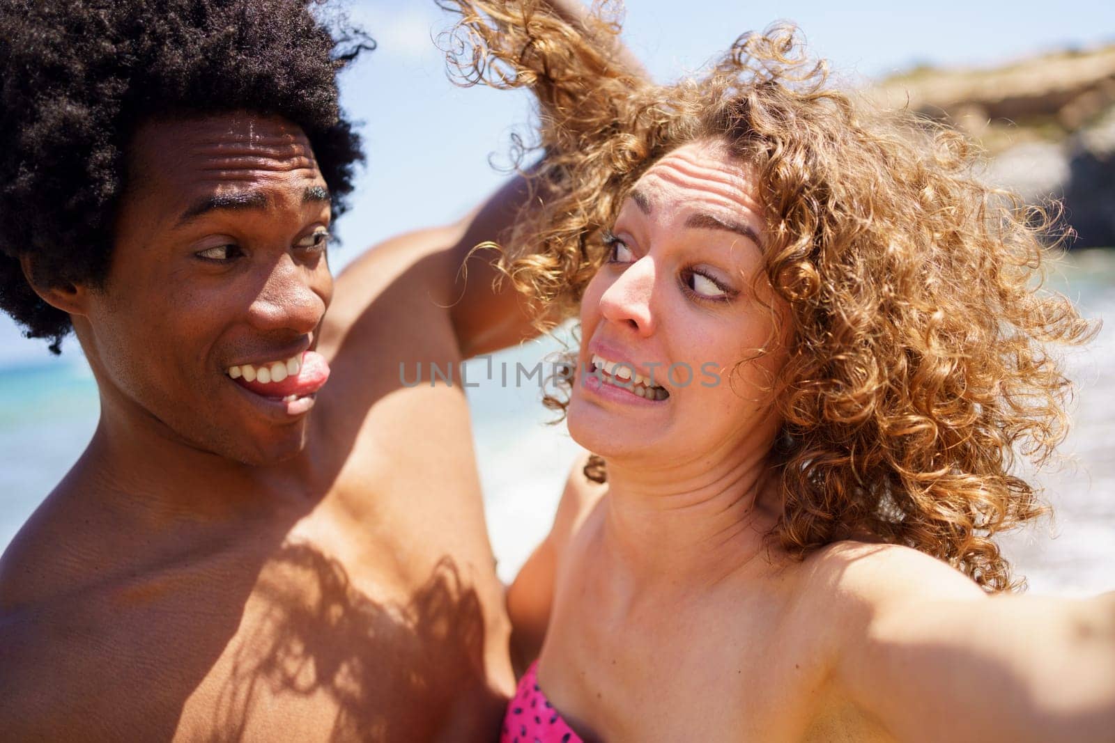 Cheerful black man with Afro hair and tongue out touching curly hair of girlfriend making funny grimace on sunny day near sea