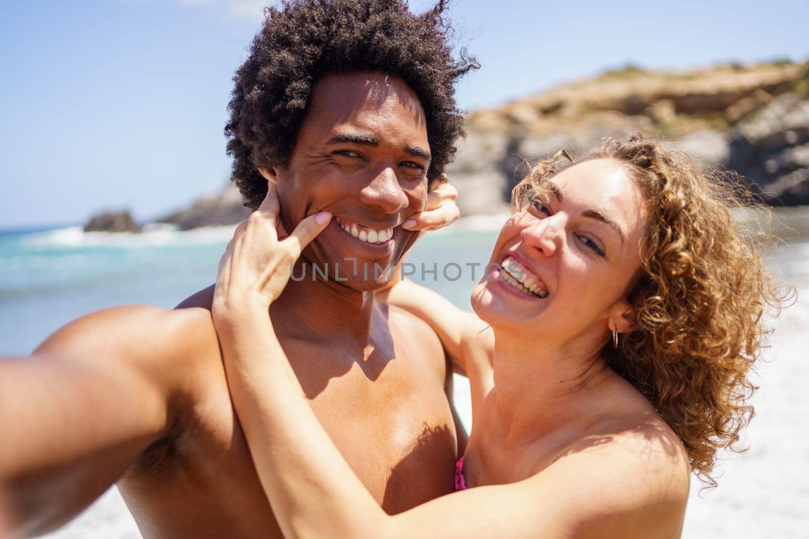 Cheerful young woman pulling smile on black boyfriend with Afro hair by stretching corners of mouth while taking selfie on beach during summer vacation