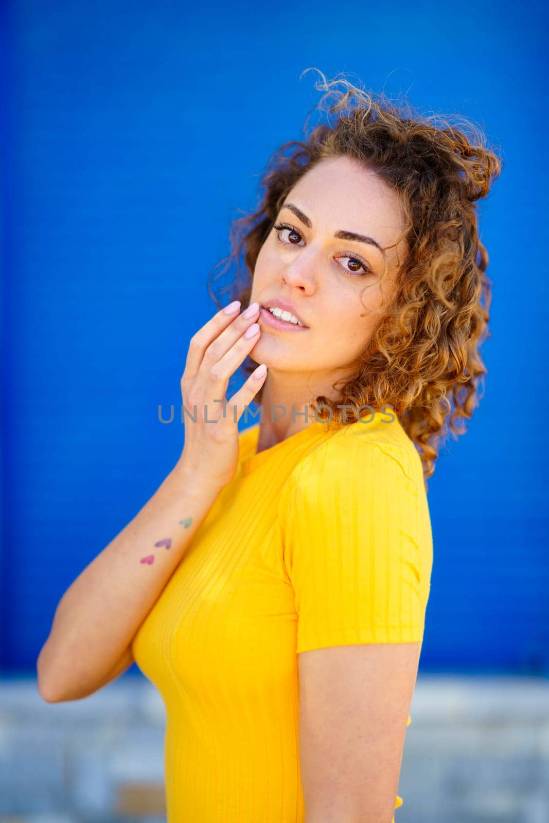 Charming young female in yellow t shirt with curly brown hair looking at camera and touching lips against blue background on blurred city street