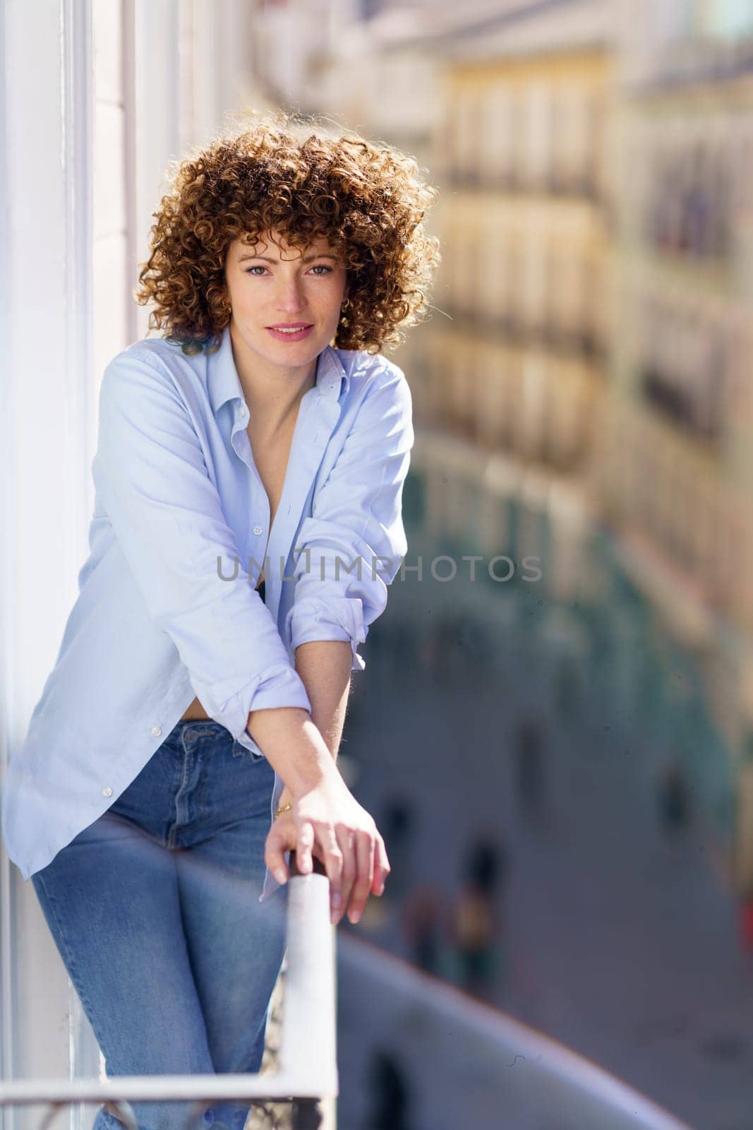 Curly haired woman in blue shirt and jeans leaning on metal railing of balcony by javiindy