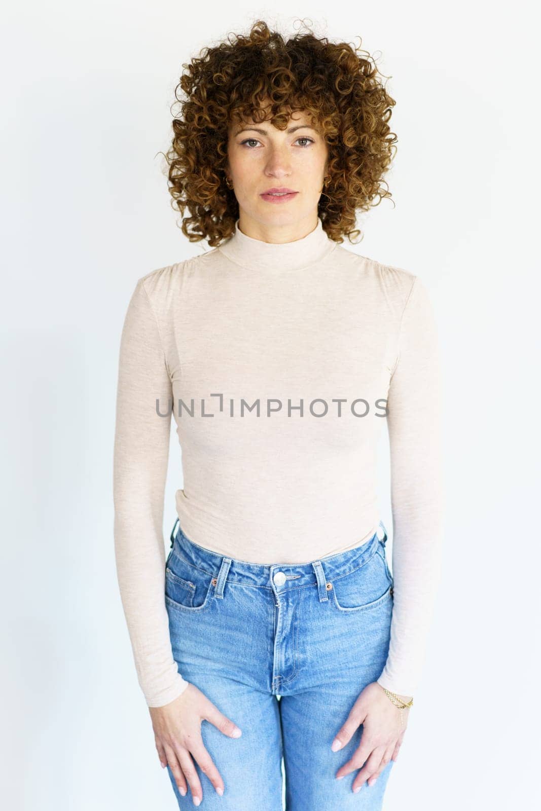Adult female in beige turtleneck and jeans with brown curly hair looking at camera while standing still with arms hanging straight on sides
