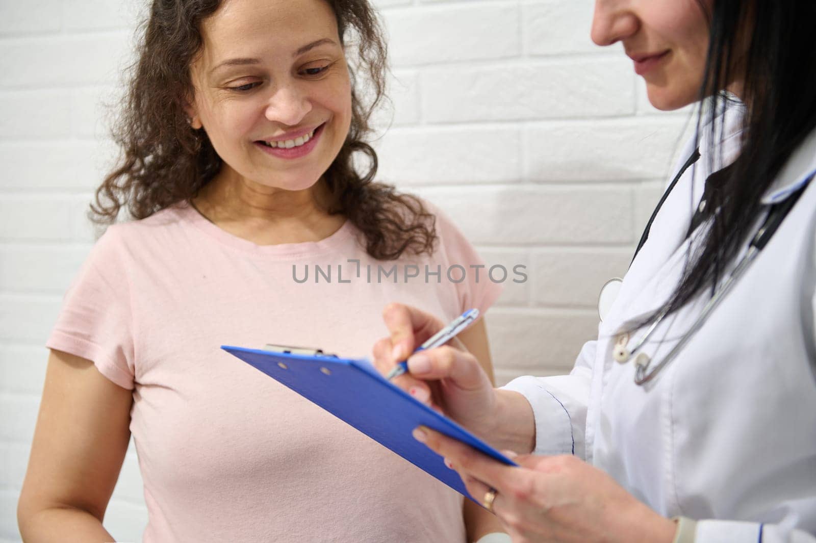 Close-up portrait of a muti ethnic adult pregnant woman in pink t-shirt, smiling when female doctor prescribing medicines. Health care. Gynecology and obstetrics. Compliance. Preganncy and maternity