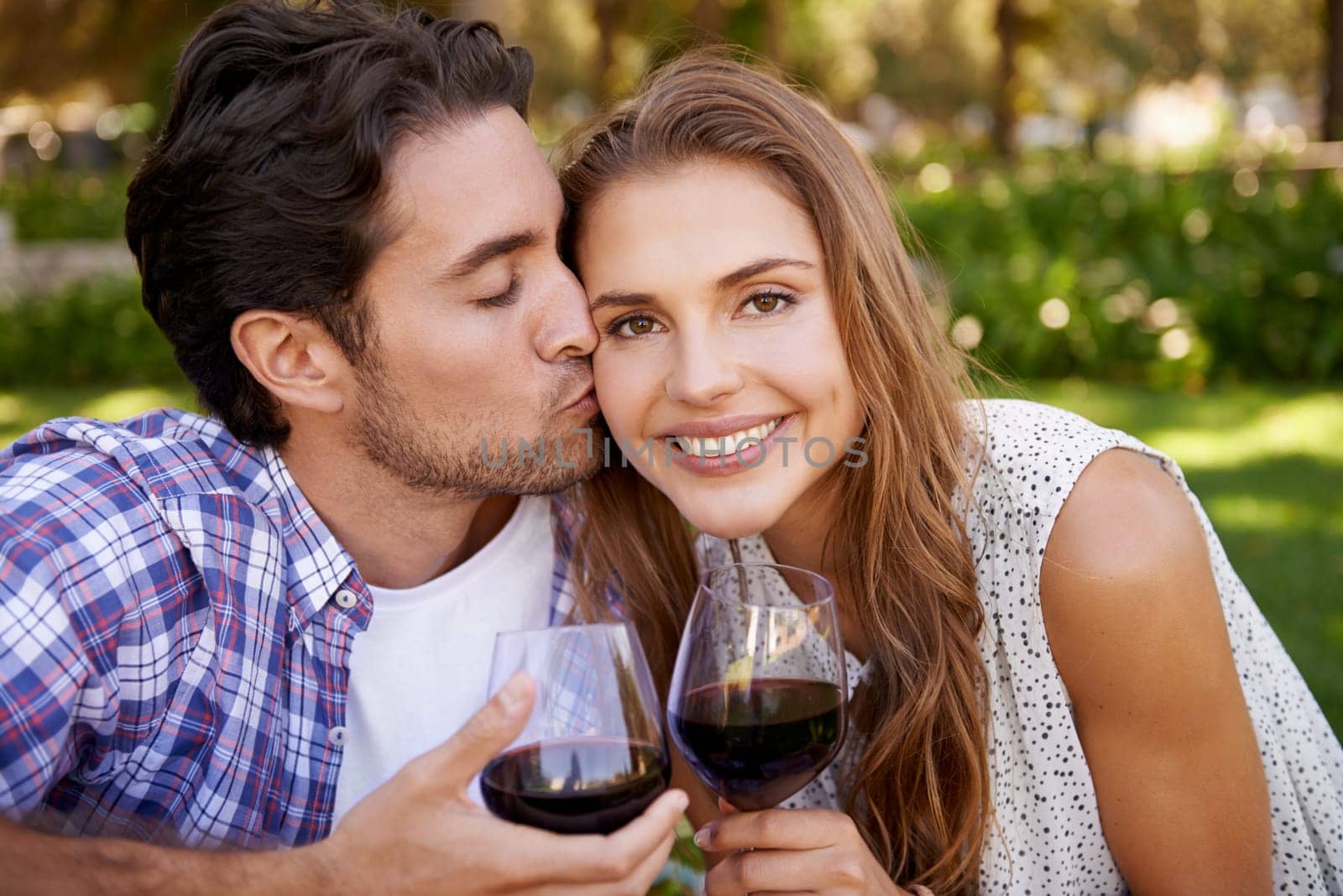 Couple portrait, wine or cheek kiss on picnic date, valentines day or romance bonding in nature, park or garden. Smile, happy woman and man with alcohol drinks glass for love anniversary celebration.