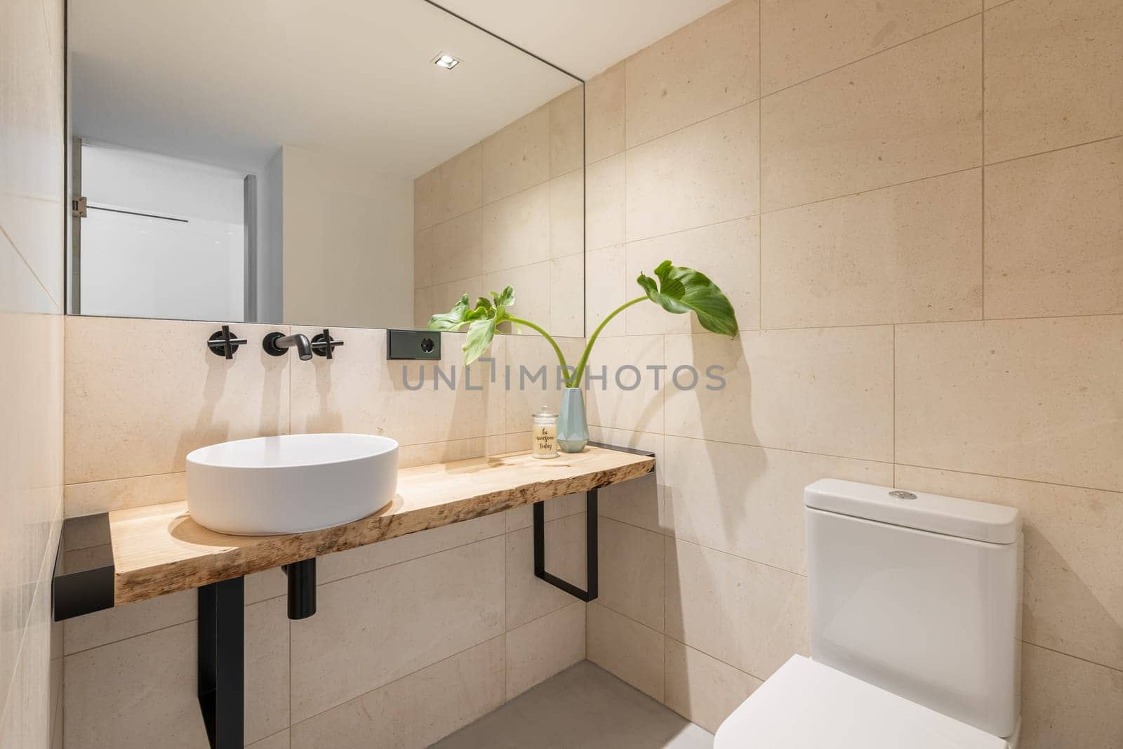 Stylish bright bathroom with beige tiles on the walls and a large mirror with a sink in a wooden countertop and a toilet. The concept of an unusual but concise shower design.