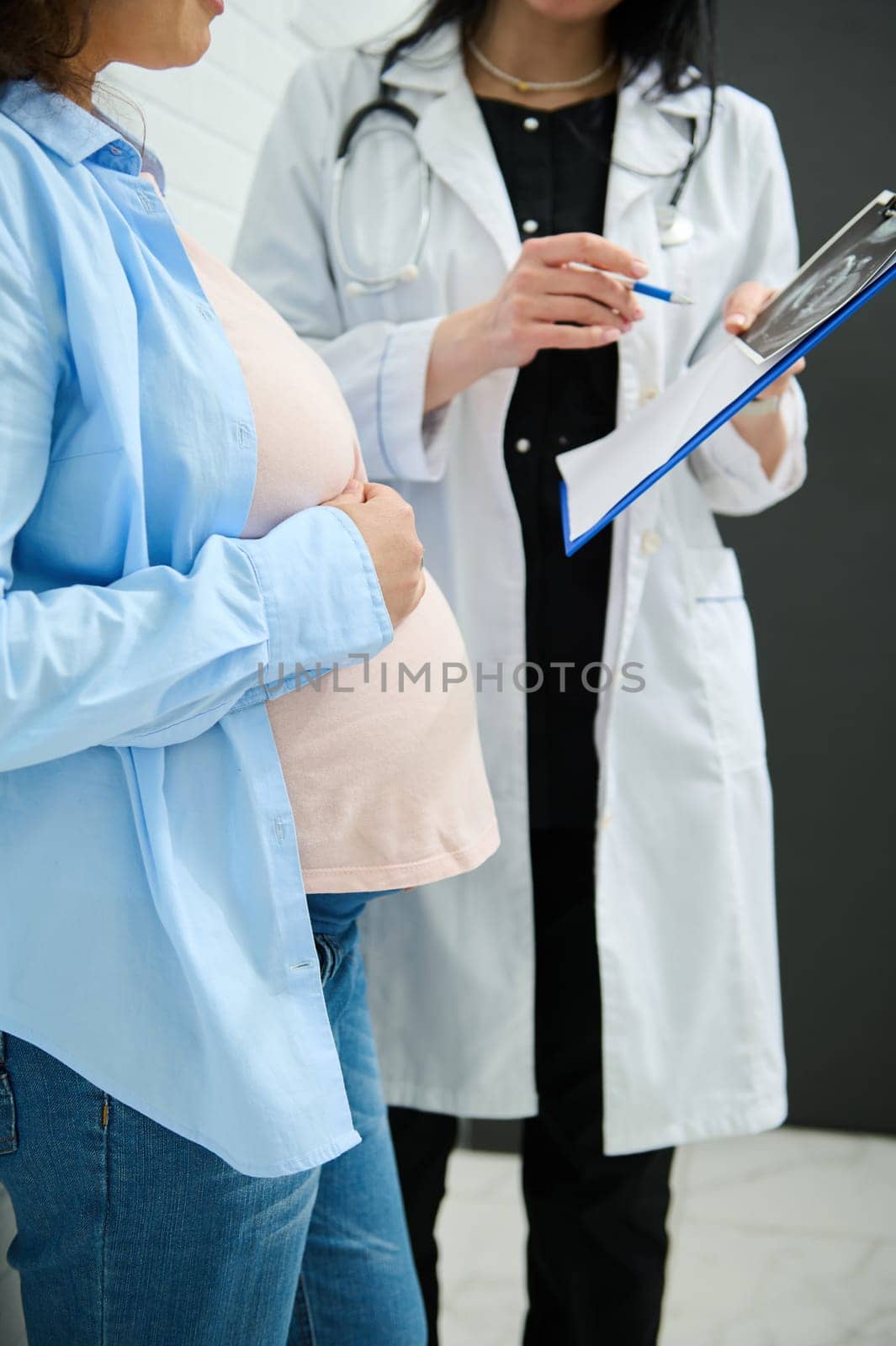 Mid-section image. Unrecognizable female doctor explaining ultrasound scan results to pregnant woman in obstetric clinic by artgf