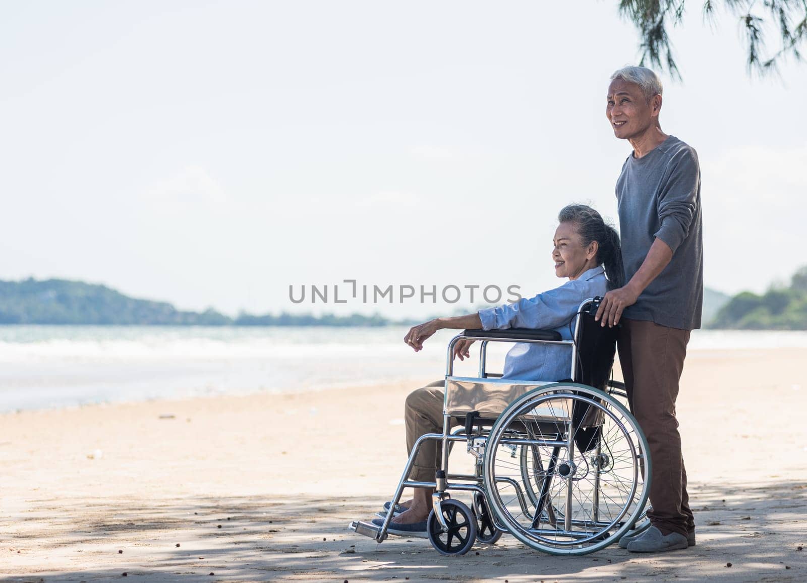 Happy Asian back elderly woman disabled sitting in wheelchair and husband is a wheelchair user on the beach together, summer vacation, Retirement couple concept