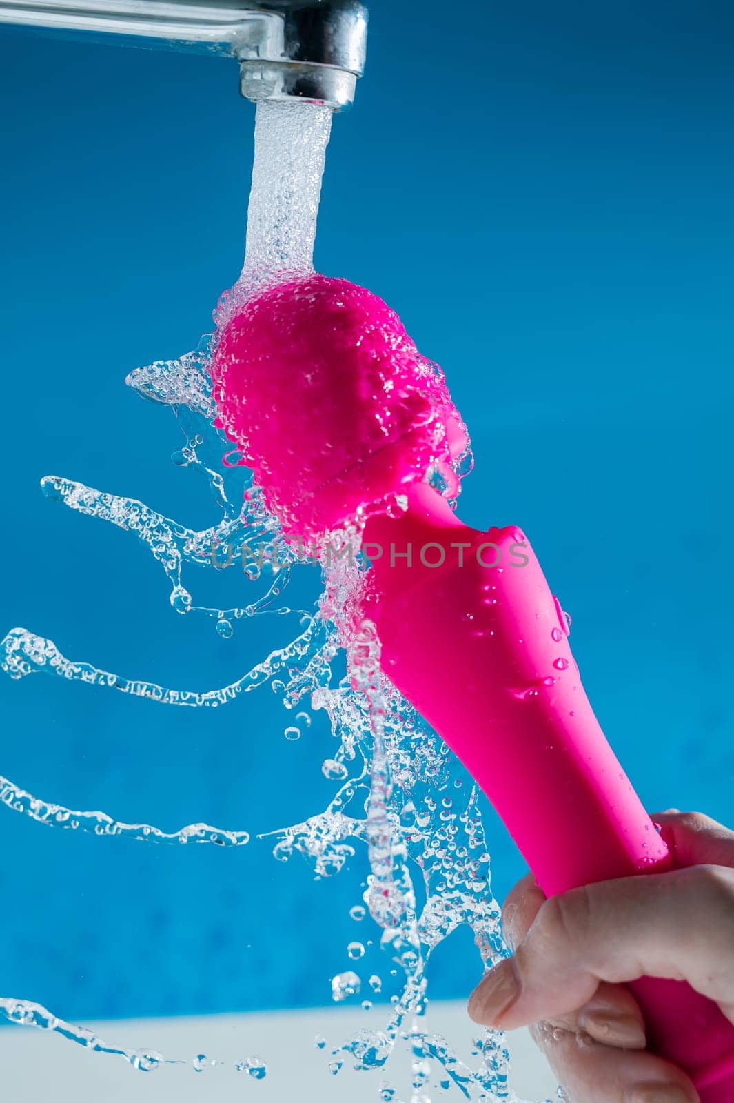 A woman washes a pink vibrator under running water on a blue background. Sex toy hygiene concept