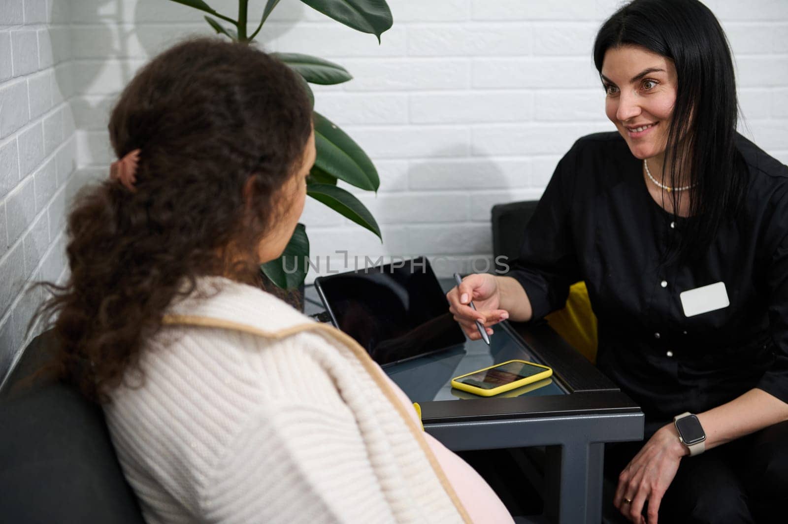 Beautiful female doctor in black uniform talking to her patient, sitting together on sofa in a modern clinic interior by artgf