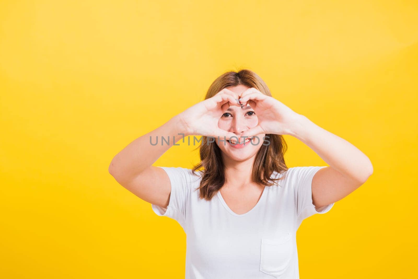 Asian Thai happy portrait beautiful cute young woman smile standing make finger heart figure symbol shape sign with two hands and looking camera, studio shot isolated yellow background with copy space