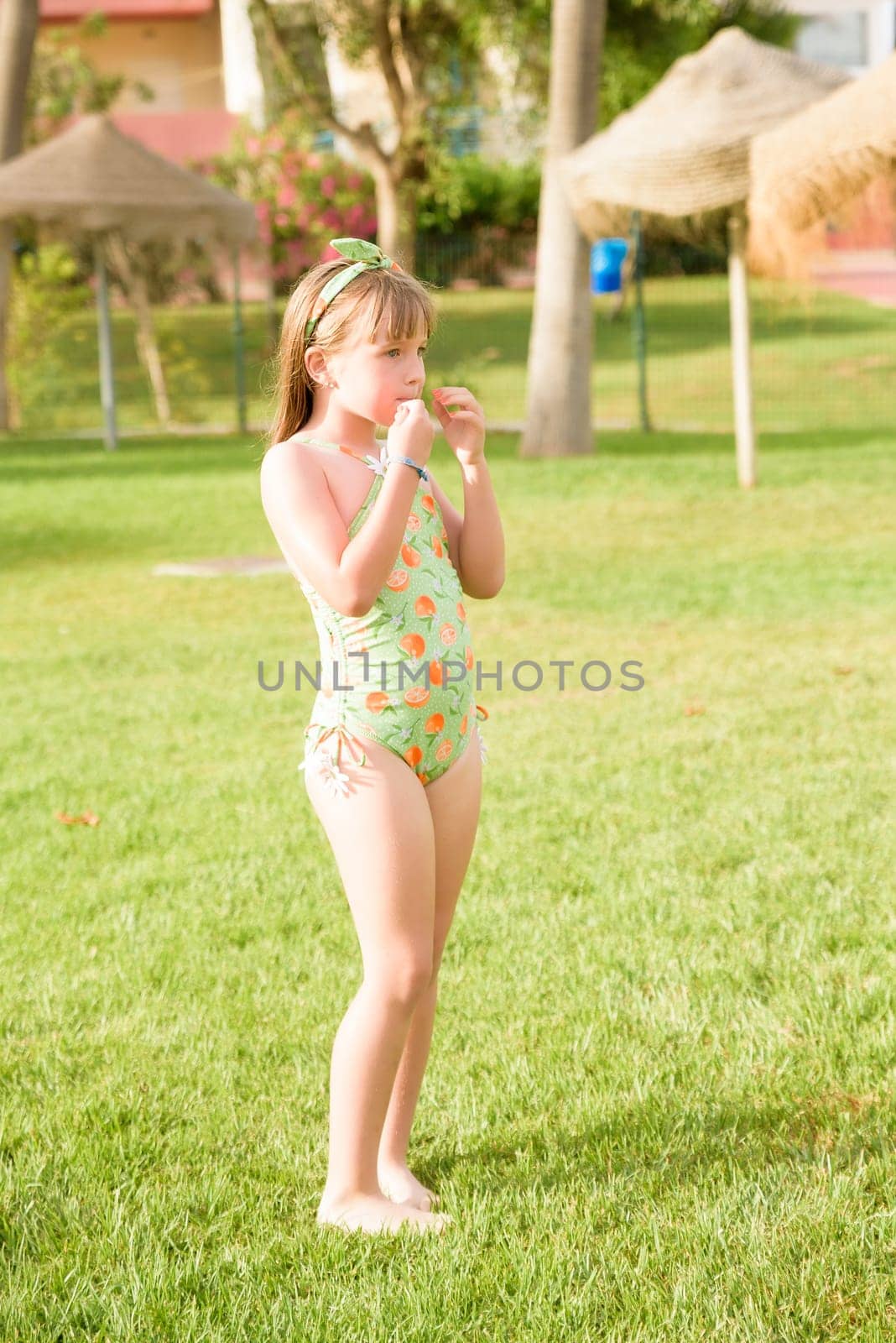 A girl in a bathing suit on the grass near the halls of the square watches something in her hands. The child has fun with the summer vacation. Family vacation by the sea.