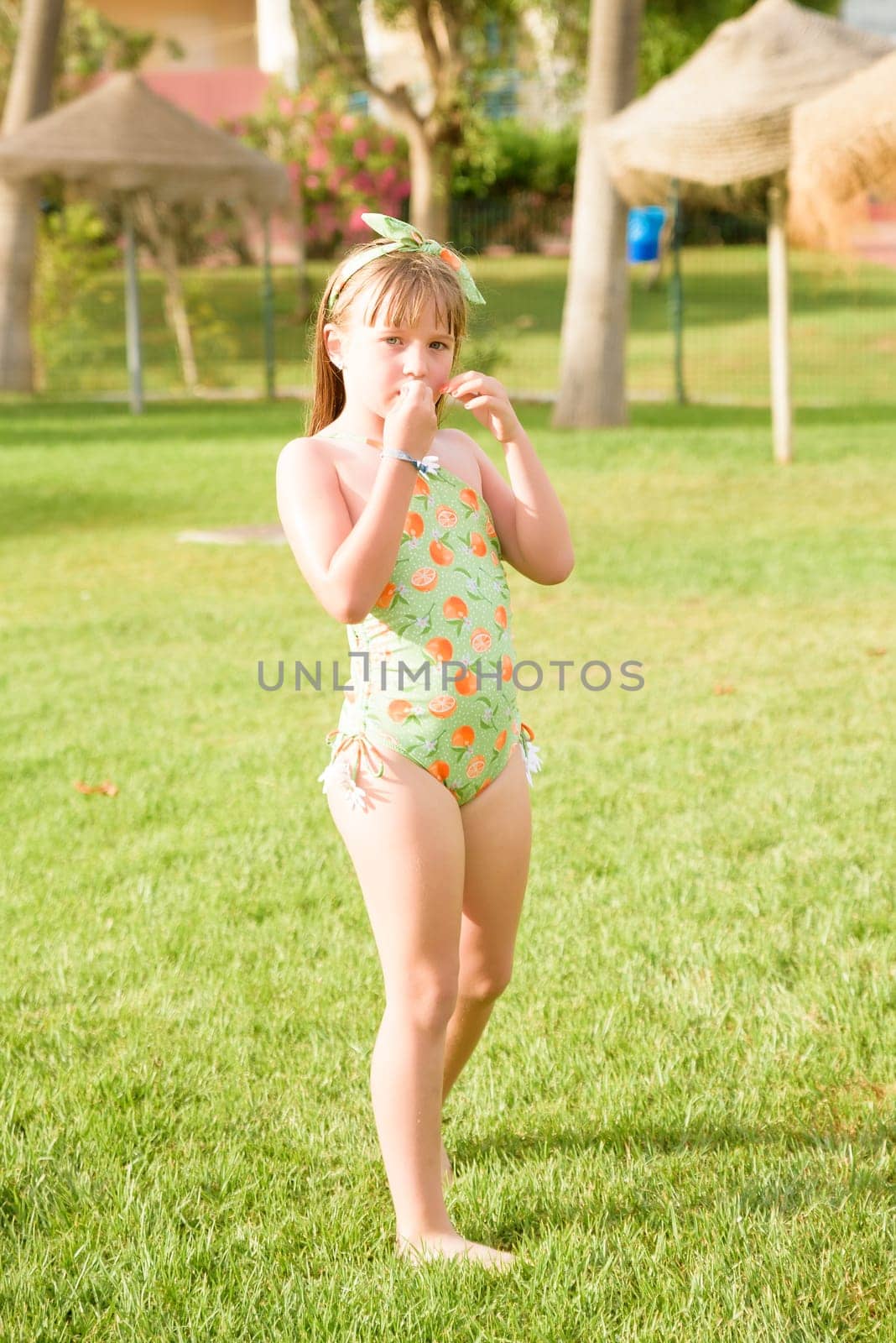 A girl in a bathing suit on the grass near the halls of the square watches something in her hands. The child has fun with the summer vacation. Family vacation by the sea.