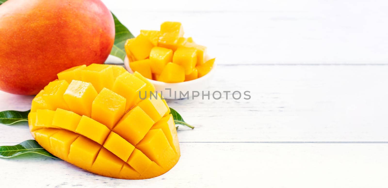 Beautiful chopped mango chunks with green leaves on bright white color timber background. Copy space, close up, macro. Tropical fruit concept.
