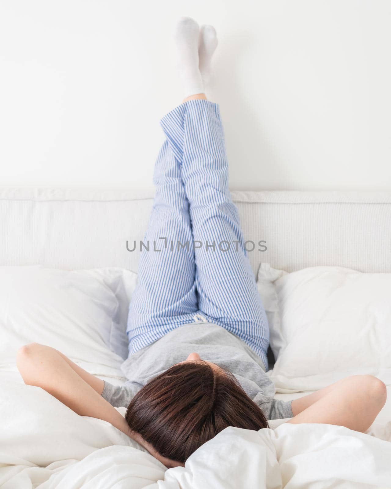 Female legs raised up high and arms under her head lying on bed in bedroom wearing pajamas by Mariakray