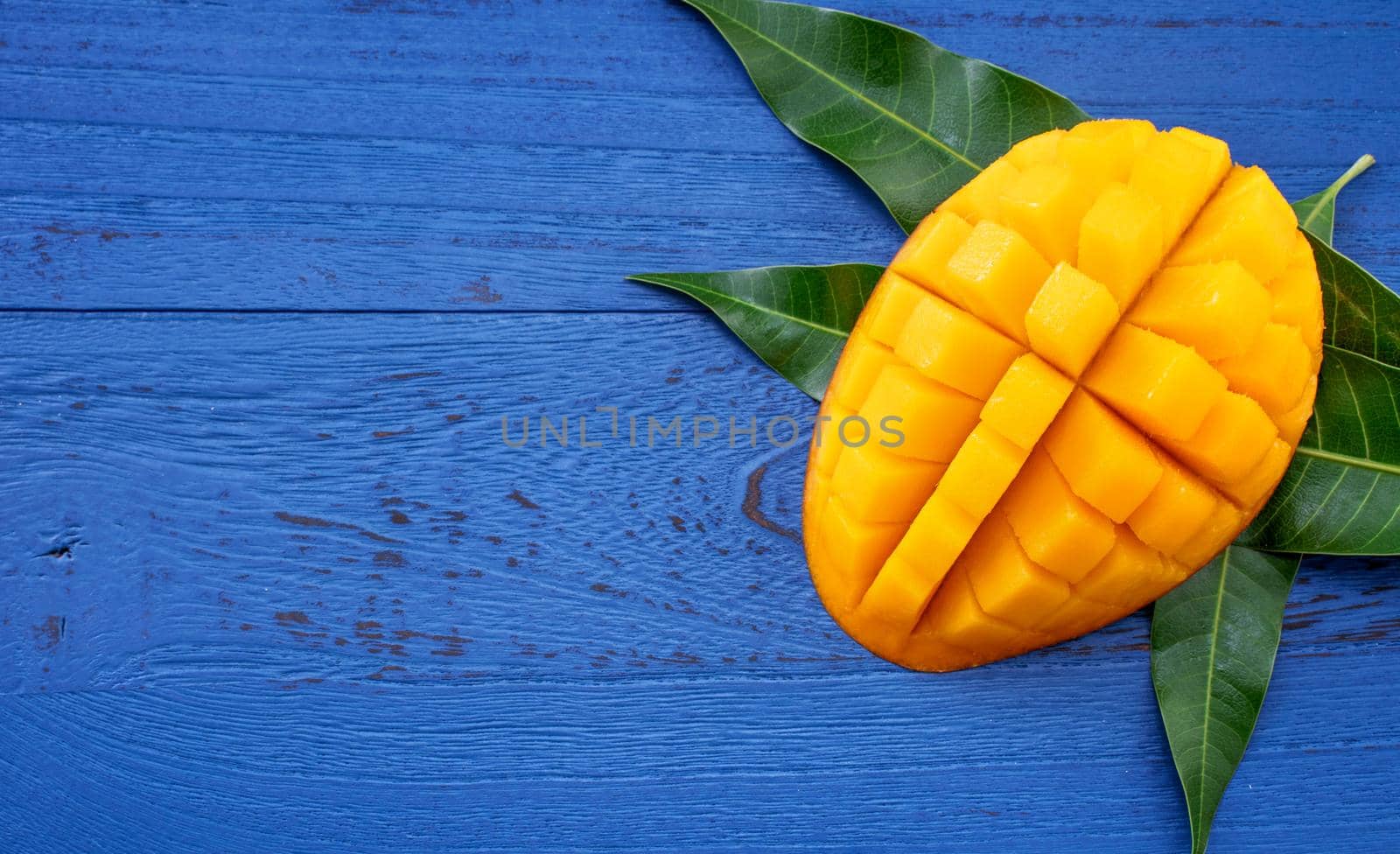 Fresh mango - beautiful chopped fruit with green leaves on dark blue timber background. Tropical fruit design concept. Flat lay. Top view. Copy space