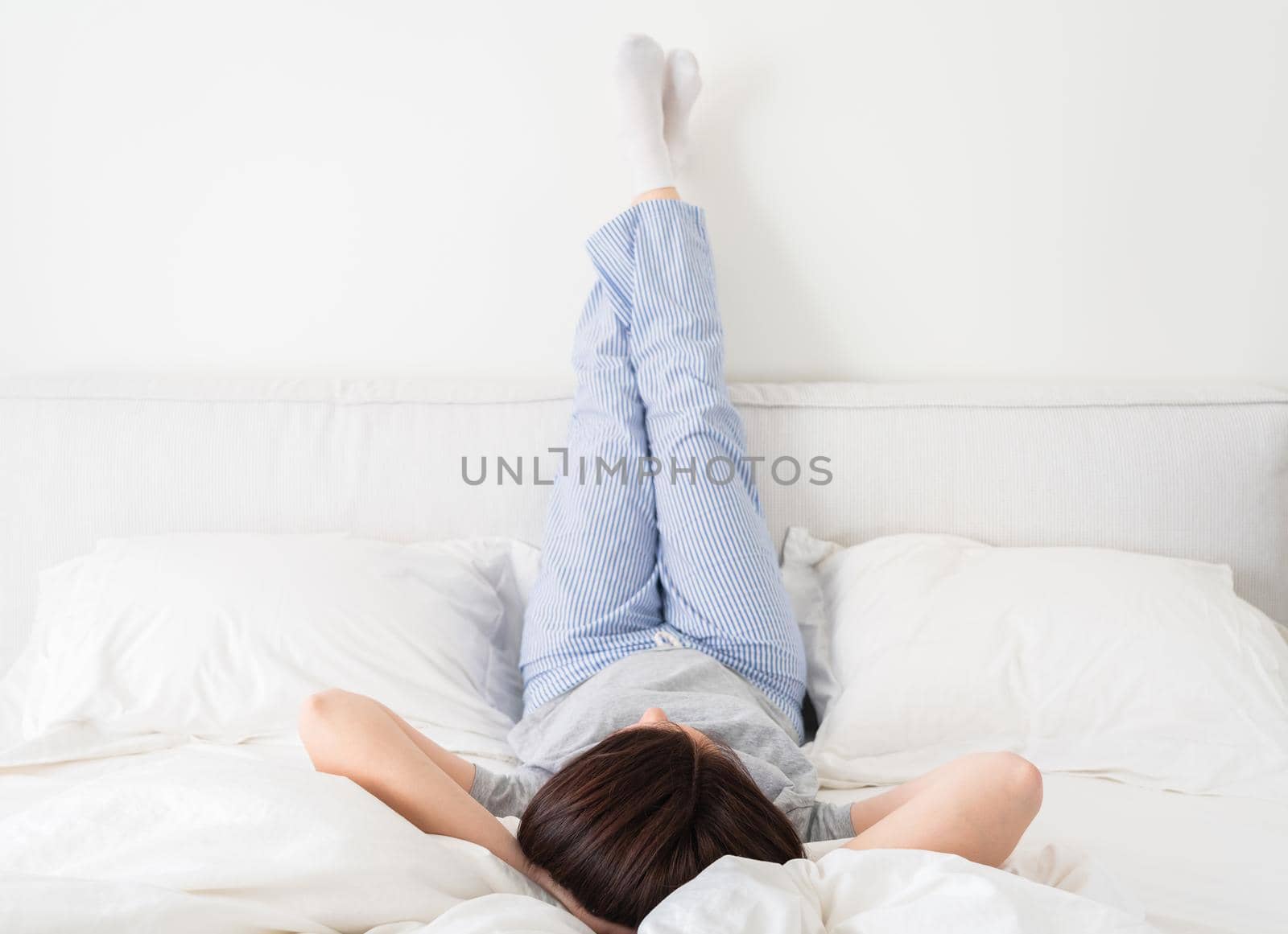 Woman with legs raised up high and arms under her head lying on bed in bedroom by Mariakray