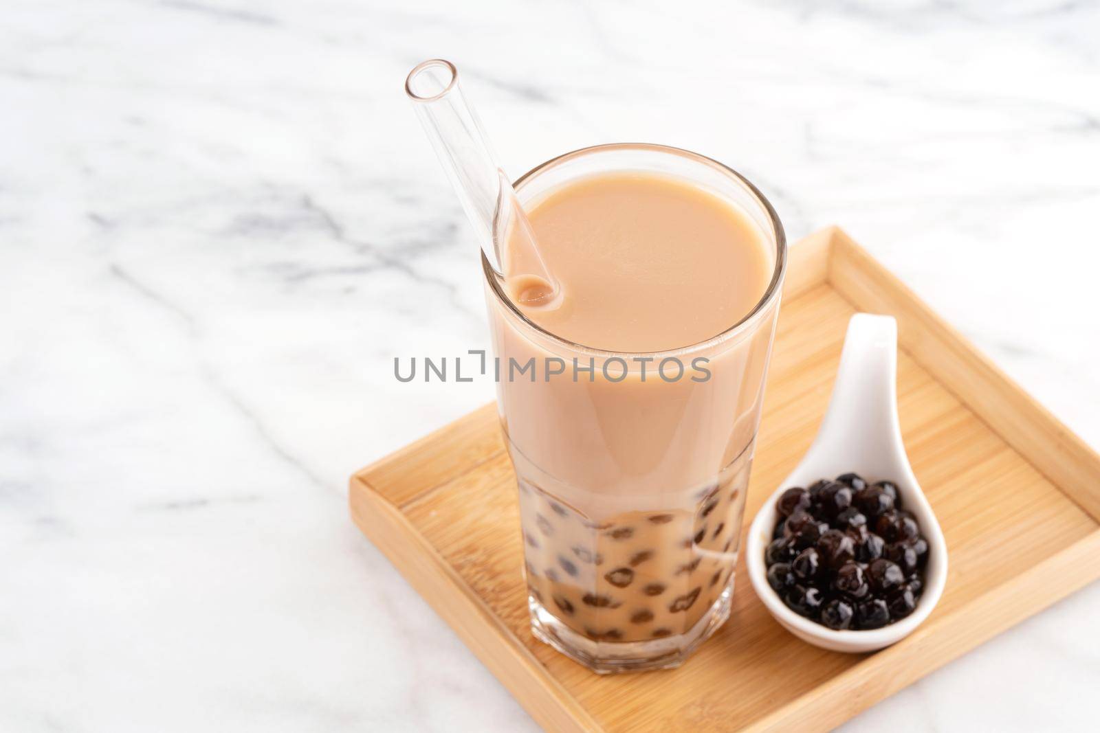 Tapioca pearl ball bubble milk tea, popular Taiwan drink, in drinking glass with straw on marble white table and wooden tray, close up, copy space.