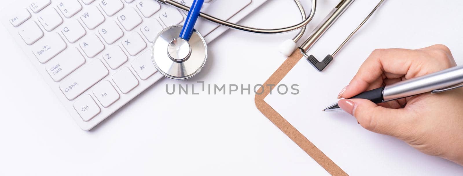 Female doctor writing a medical record case over clipboard on white working table with stethoscope, computer keyboard. Top view, flat lay, copy space