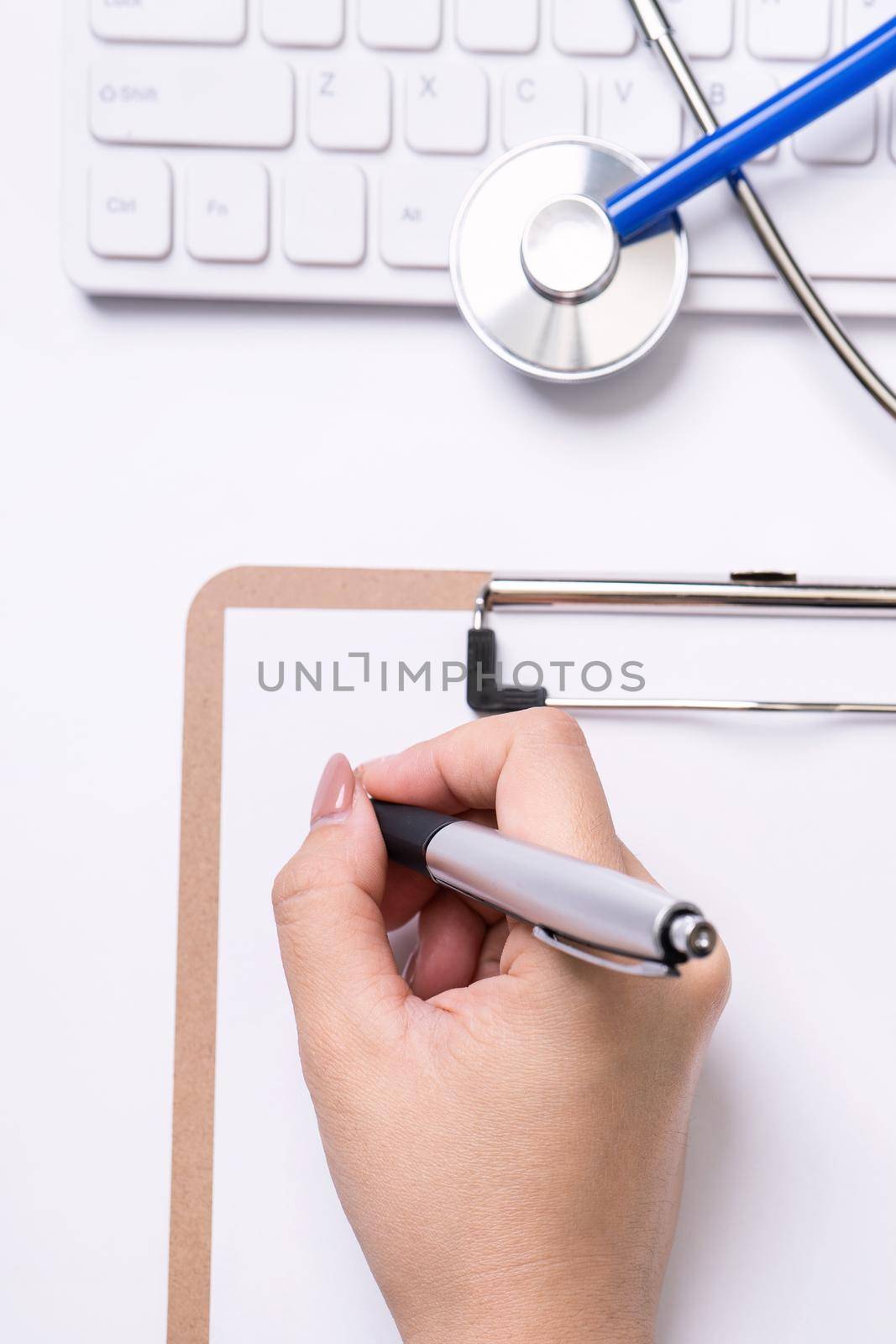 Female doctor writing a medical record case over clipboard on white working table with stethoscope, computer keyboard. Top view, flat lay, copy space