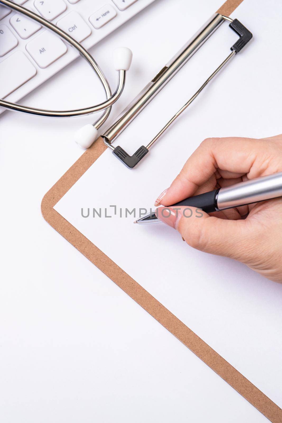 Female doctor writing a medical record case over clipboard on white working table with stethoscope, computer keyboard. Top view, flat lay, copy space