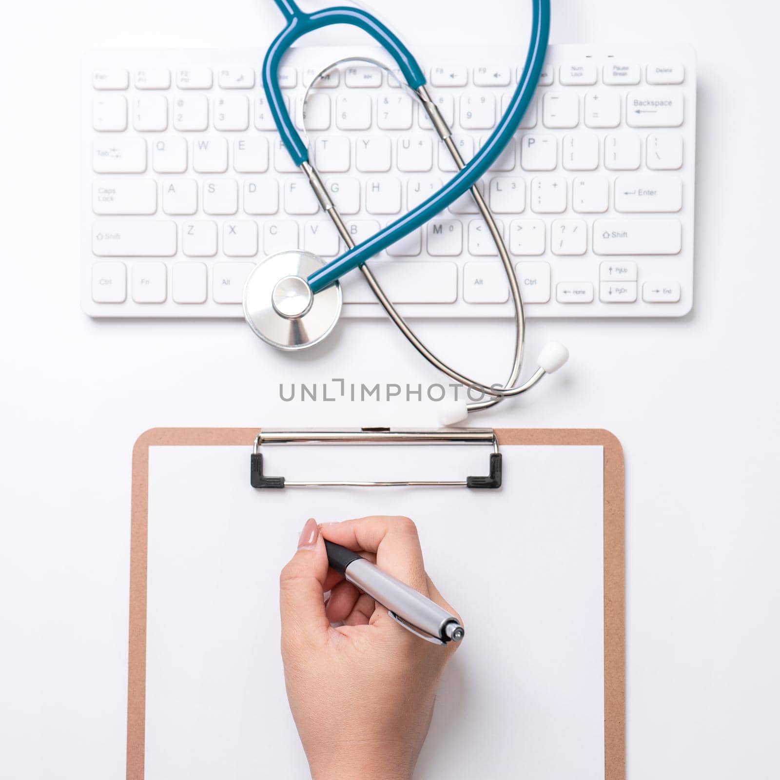 Female doctor writing a medical record case over clipboard on white working table with stethoscope, computer keyboard. Top view, flat lay, copy space