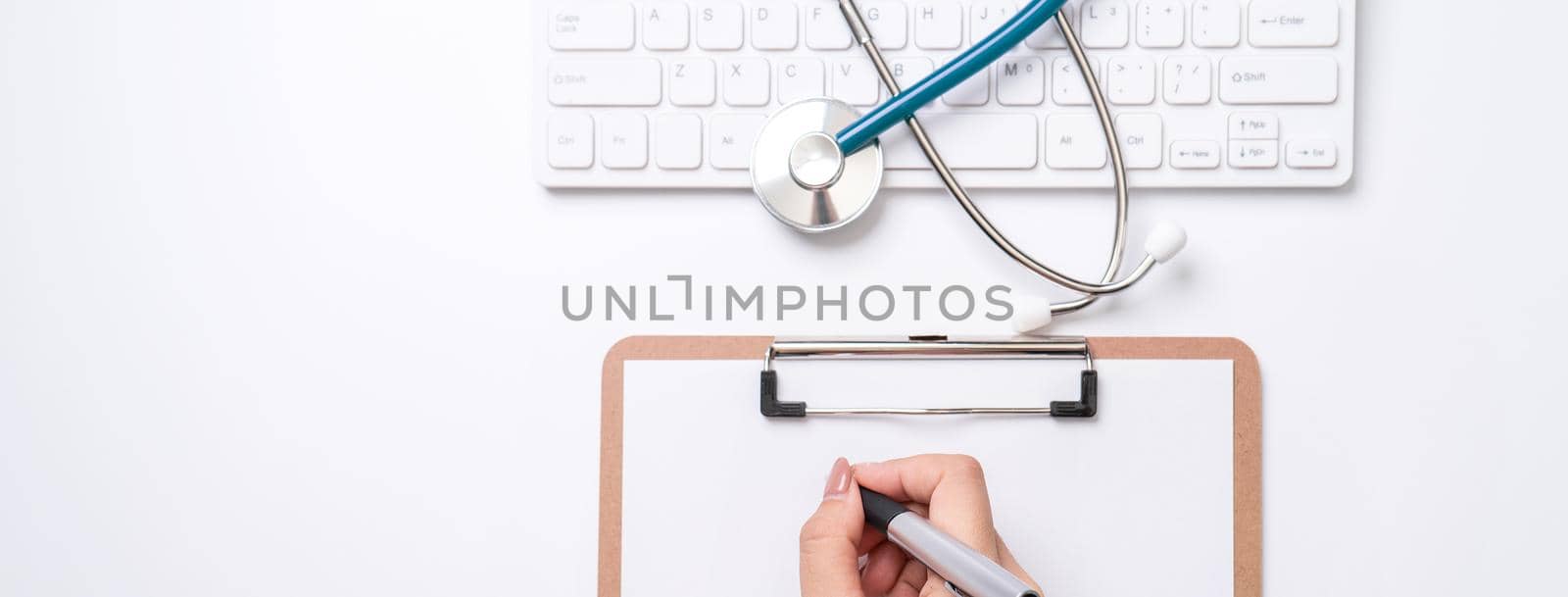 Female doctor writing a medical record case over clipboard on white working table with stethoscope, computer keyboard. Top view, flat lay, copy space