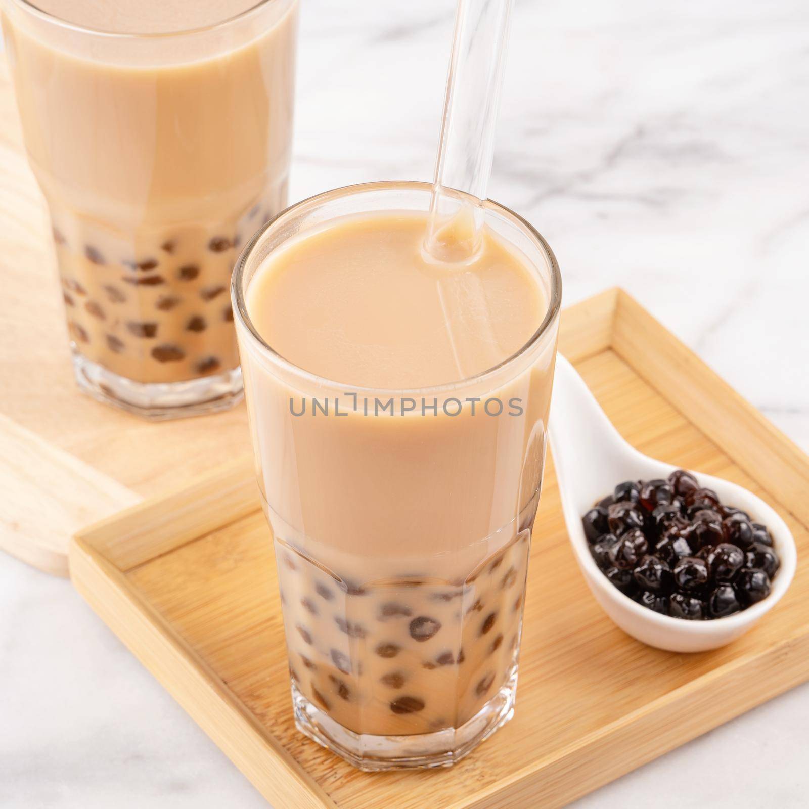 Tapioca pearl ball bubble milk tea, popular Taiwan drink, in drinking glass with straw on marble white table and wooden tray, close up, copy space.