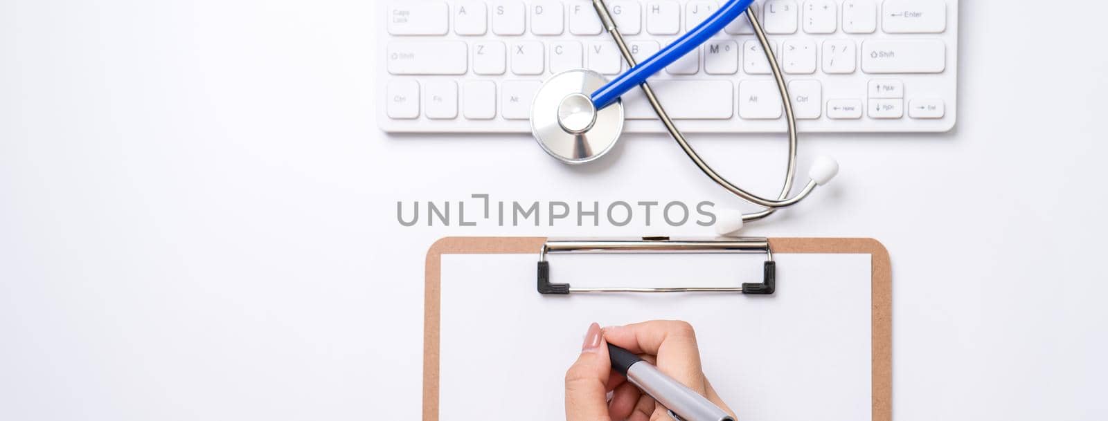 Female doctor writing a medical record case over clipboard on white working table with stethoscope, computer keyboard. Top view, flat lay, copy space by ROMIXIMAGE