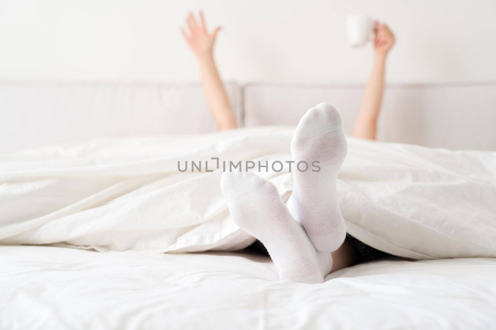 Female hand holding cup of coffee from under a blanket in bed. Woman waking up in the morning by Mariakray