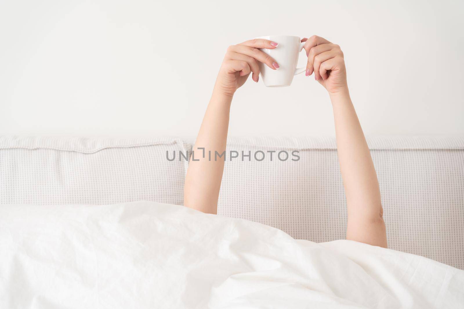 Female hand holding cup of coffee from under a blanket in bed. Woman waking up in the morning.