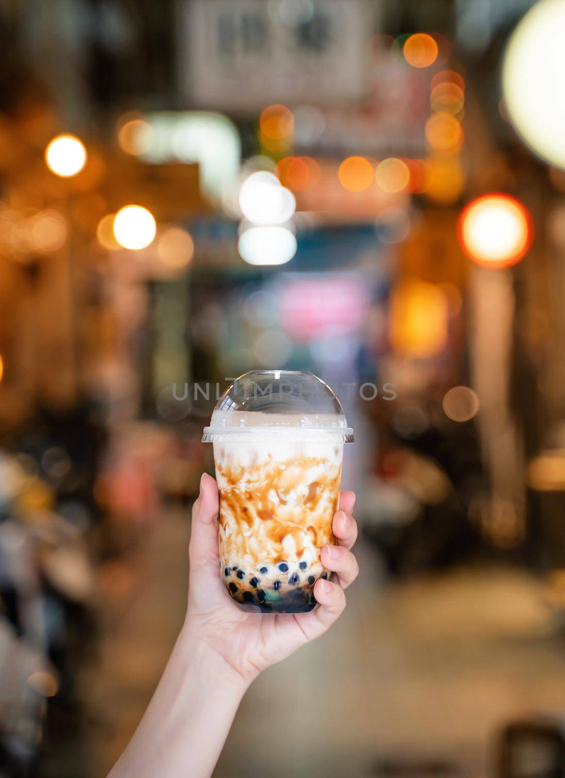 Young girl is holding and showing a cup of brown sugar flavored tapioca pearl bubble milk tea in night market of Taiwan background, close up, bokeh